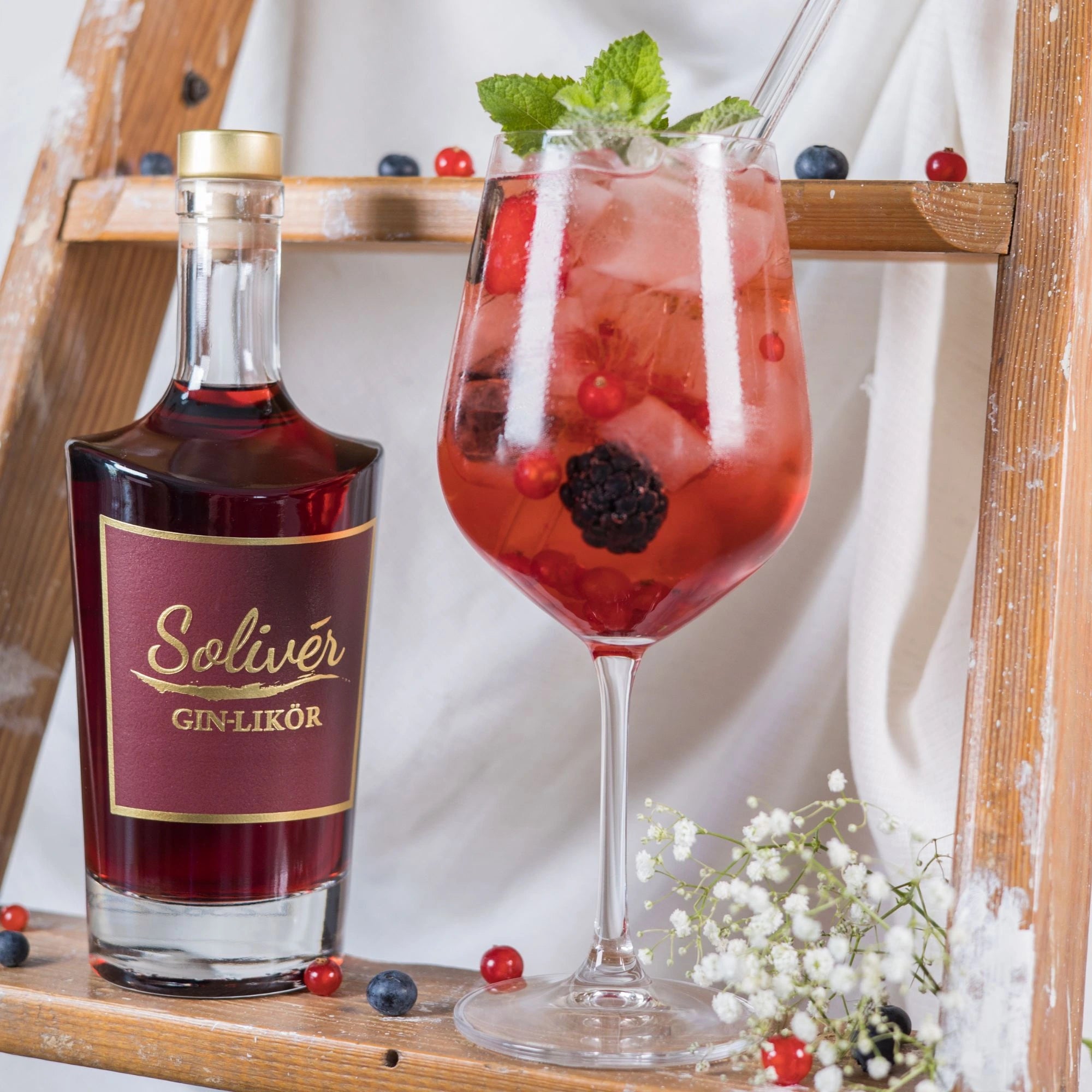 Elegante Solivér Brombeer Gin-Likör Flasche neben einem Aperitif im Weinglas mit Eiswürfeln, Beeren und Minze, dekorativ inszeniert auf einer Holzleiter.