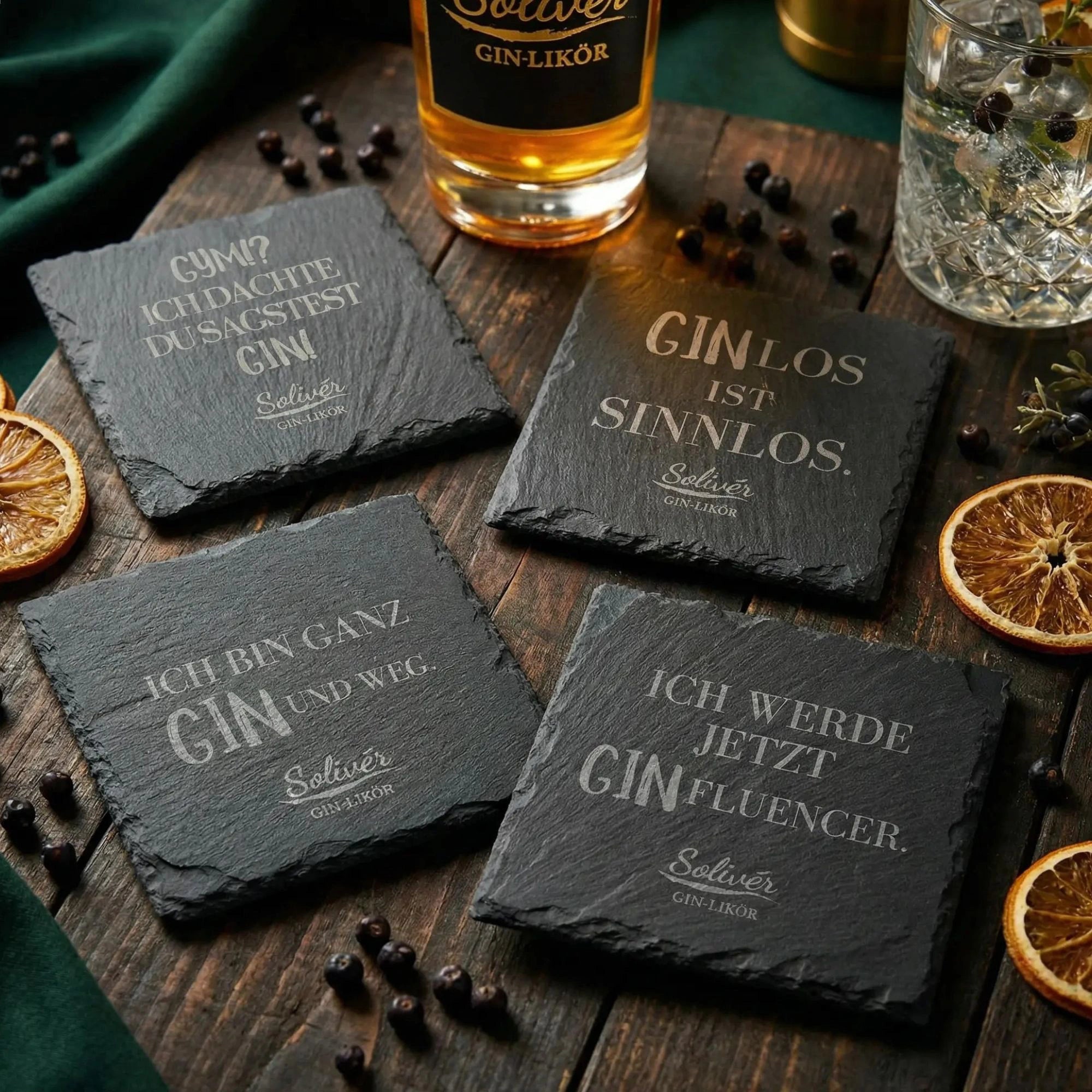 Vier schwarze Schiefer-Untersetzer mit humorvollen Gin-Sprüchen von Solivér Gin-Likör auf Holztisch, dekoriert mit Wacholderbeeren, getrockneten Orangenscheiben und Barzubehör.