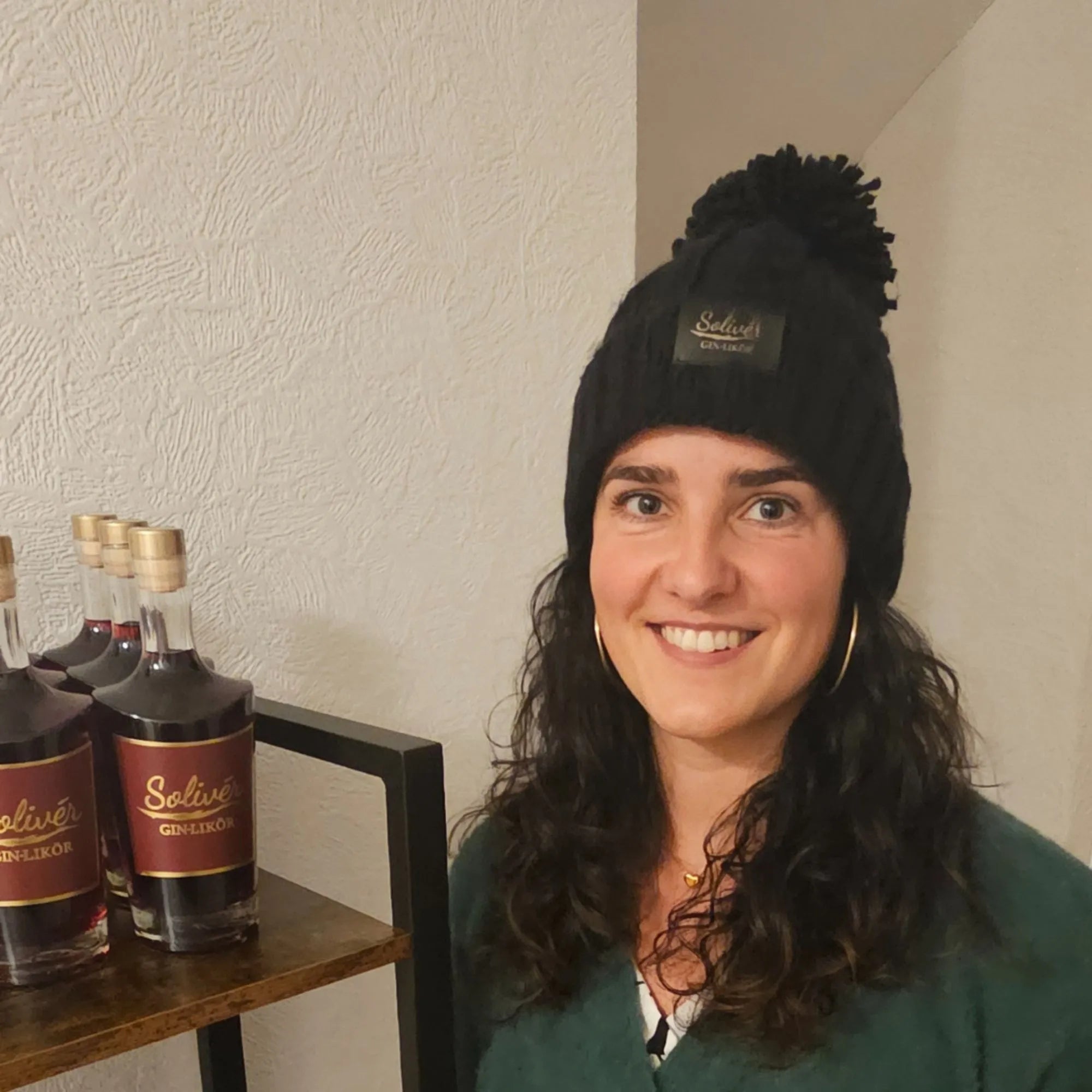 Frau mit schwarzer Solivér Beani mit Bommel steht lächelnd neben einem Regal mit mehreren Flaschen Solivér Brombeer Gin-Likör.