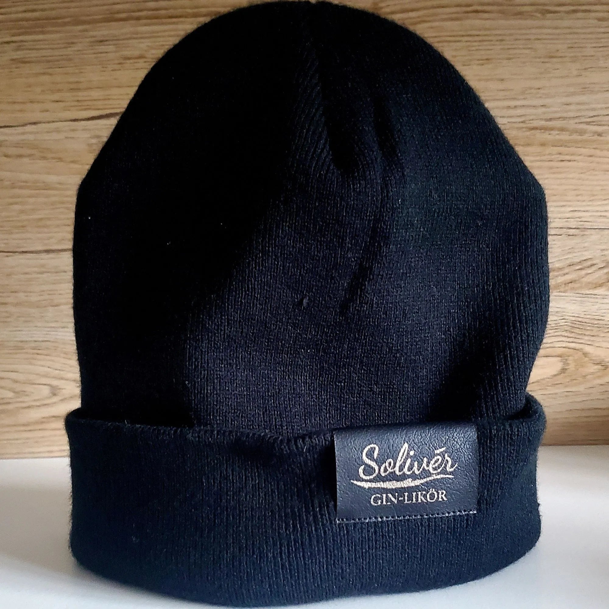 Schwarze Solivér Beani mit aufgenähtem Leder-Label auf weißem Untergrund vor hellem Holz-Hintergrund.