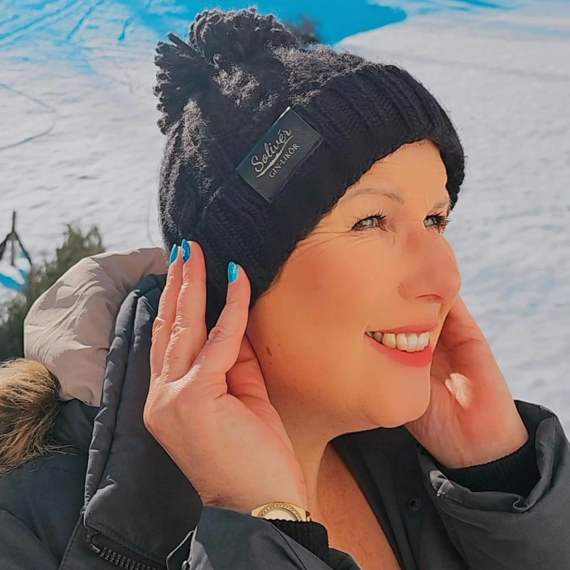 Frau lächelt in die Wintersonne und trägt eine schwarze Solivér Beani mit Bommel, kombiniert mit warmer Winterjacke.