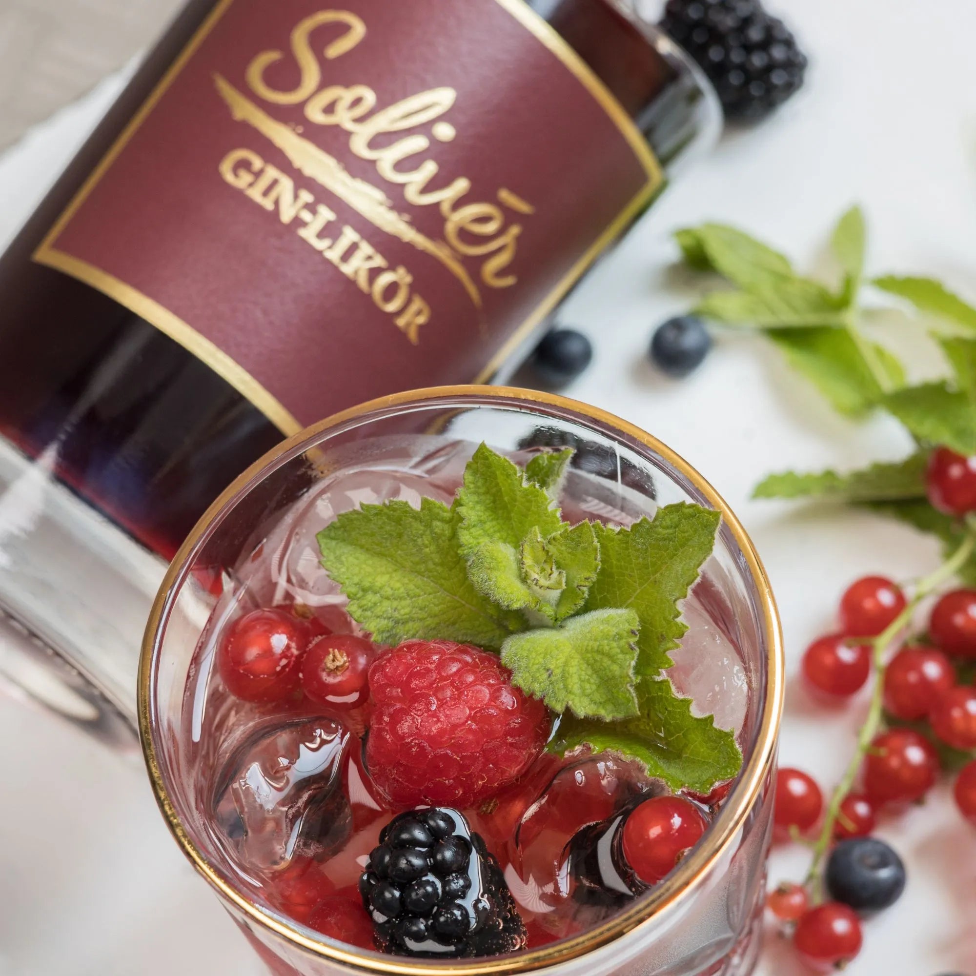 Solivér Brombeer Gin-Likör im Glas als fruchtigen Cocktail auf Eis mit frischen Beeren und Minze, daneben liegende Flasche als stilvoller Hingucker.