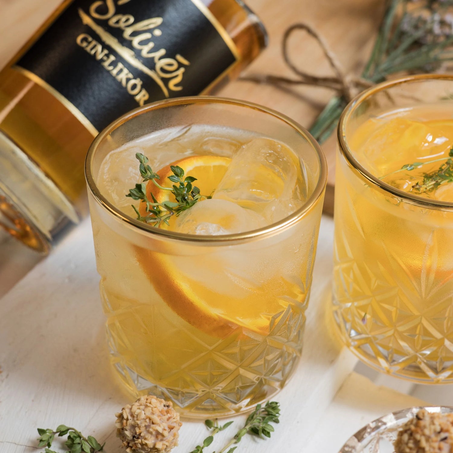 Zwei Gläser mit Solivér Goldapfel Gin-Likör auf Eis, garniert mit Orangenscheiben und frischem Thymian, daneben die passende Flasche und Dekoration aus Kräutern und Pralinen.