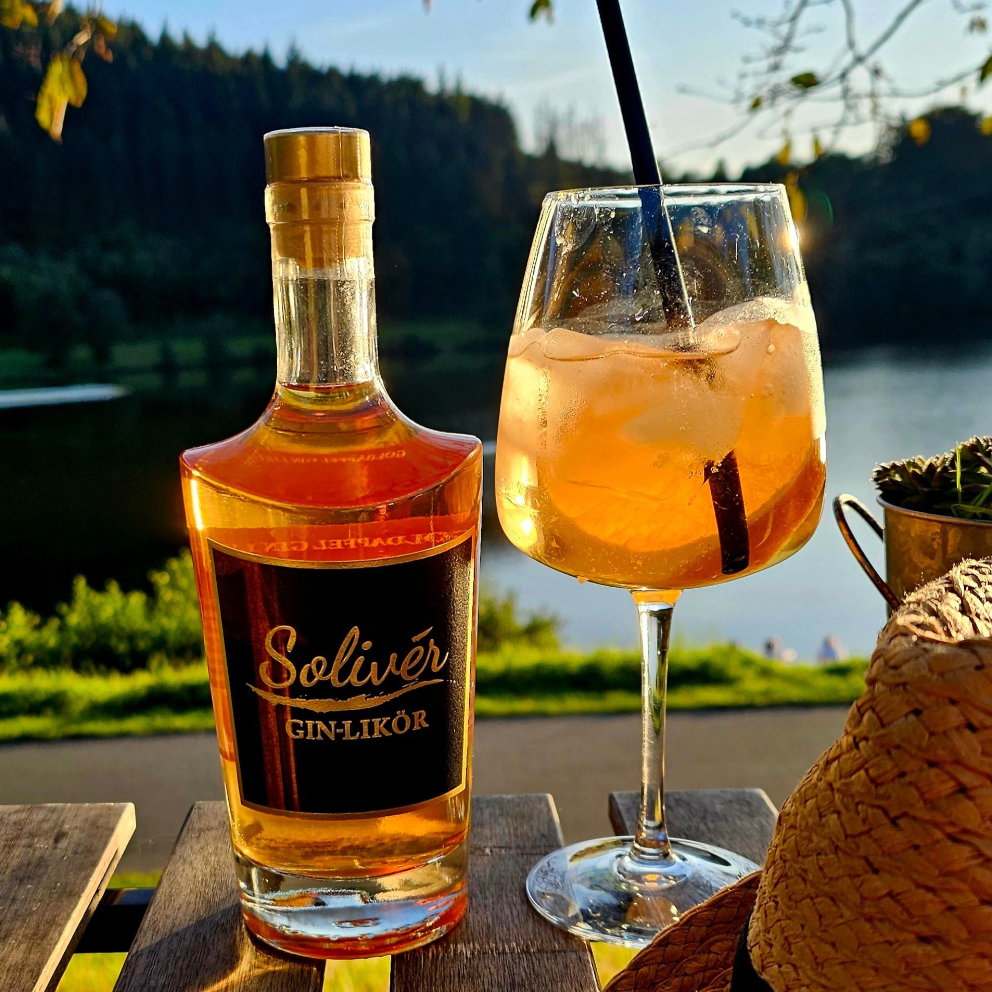 Flasche Solivér Goldapfel Gin-Likör und ein Cocktailglas mit Eiswürfeln im Sonnenlicht, aufgenommen im Freien vor einem See mit Waldkulisse.