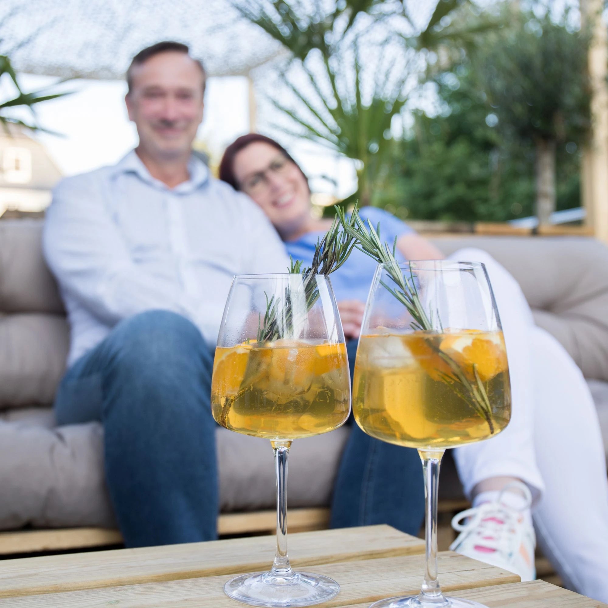 zwei gläser solivér gin-cocktail mit rosmarin vor einem paar auf einem loungesofa im garten