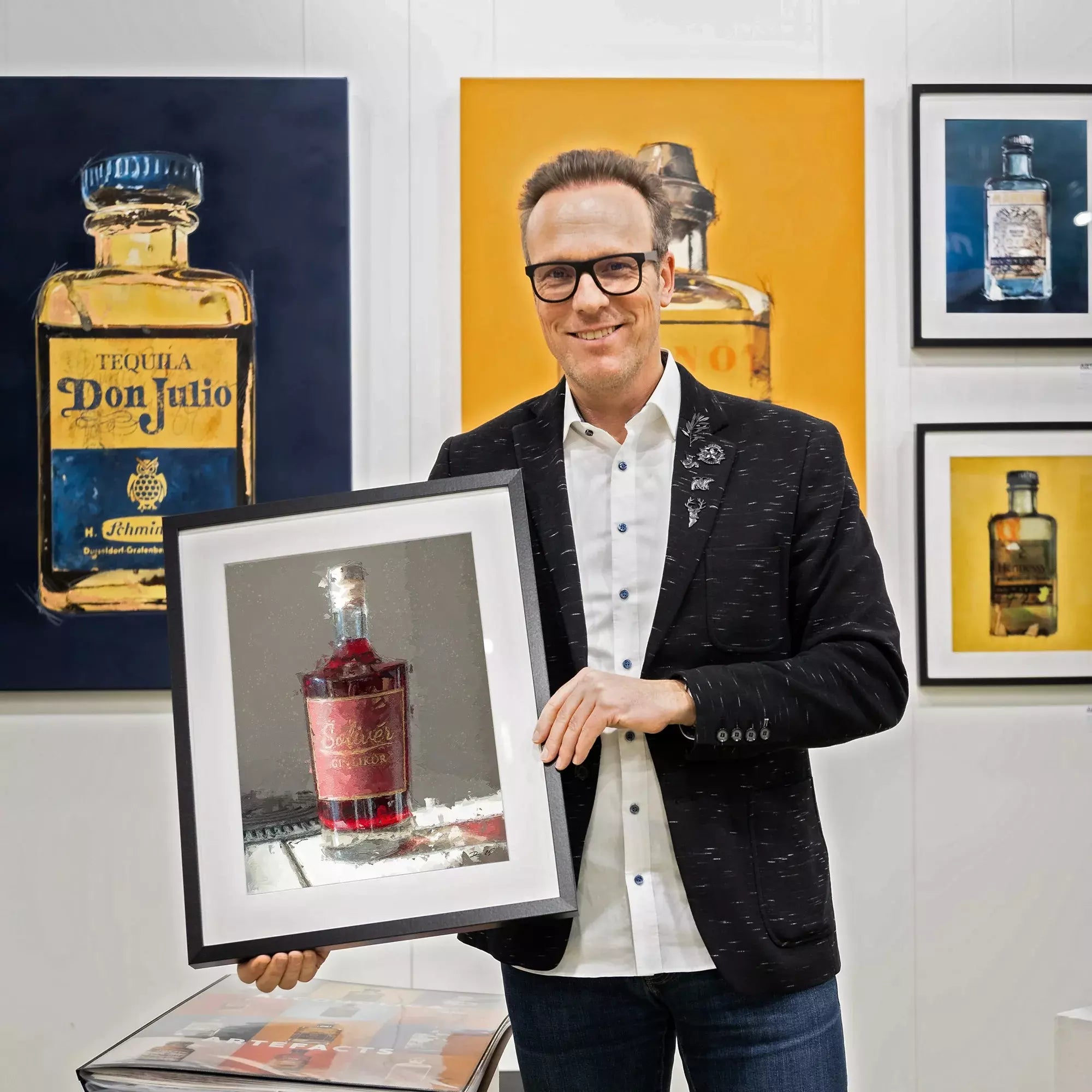 Ein Mann im Sakko hält ein gerahmtes Bild einer Solivér Ginlikör Flasche mit rotem Etikett in den Händen. Im Hintergrund hängen weitere farbige Bilder von Spirituosenflaschen an der Wand.