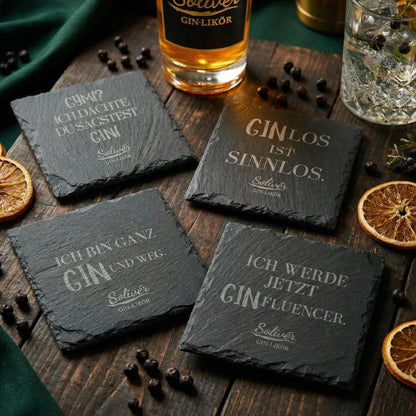 Vier schwarze Schiefer-Untersetzer mit humorvollen Gin-Sprüchen von Solivér Gin-Likör auf Holztisch, dekoriert mit Wacholderbeeren, getrockneten Orangenscheiben und Barzubehör.
