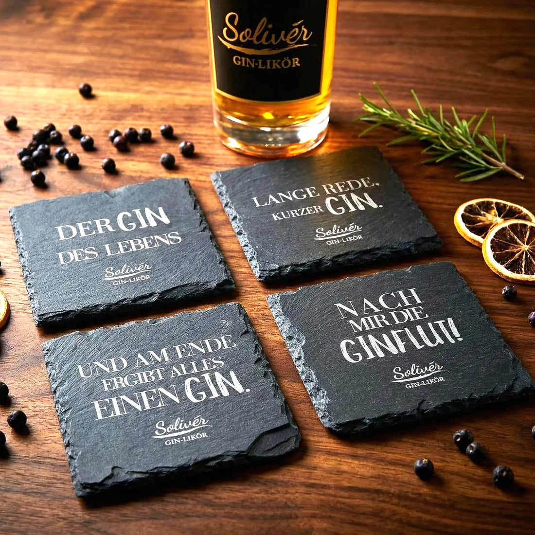 Vier schwarze Schiefer-Untersetzer mit humorvollen Gin-Sprüchen von Solivér Gin-Likör auf Holztisch, dekoriert mit Wacholderbeeren, Rosmarin und getrockneten Orangenscheiben.