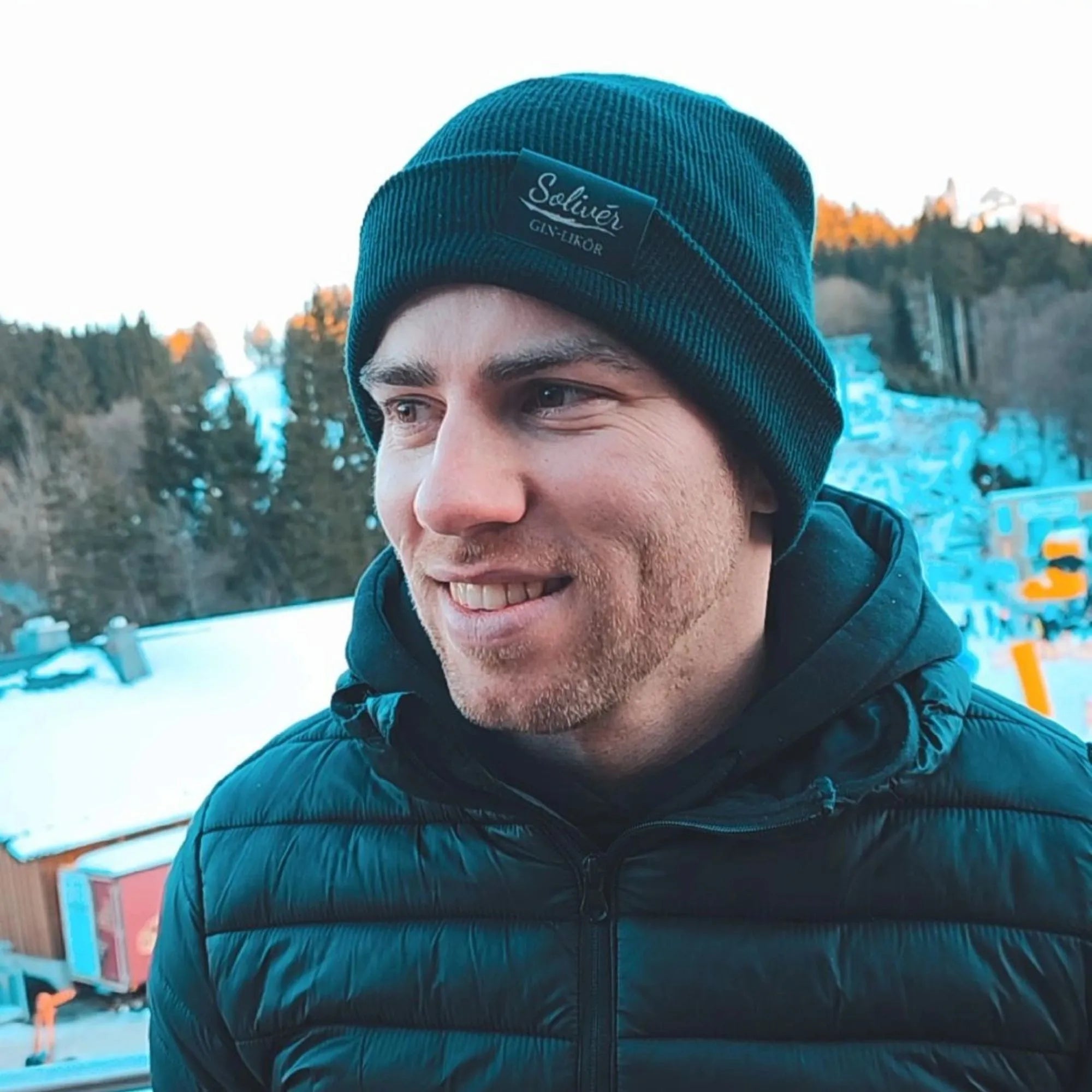 Mann mit schwarzer Solivér Beani und Winterjacke lächelt im verschneiten Outdoor-Setting mit Wald und Hütten im Hintergrund.